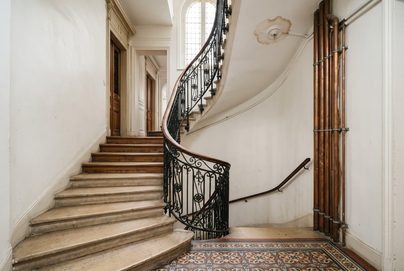 Cage d’escalier d’immeuble ancien avec canalisations apparentes le long du mur et tache d’humidité au plafond du palier