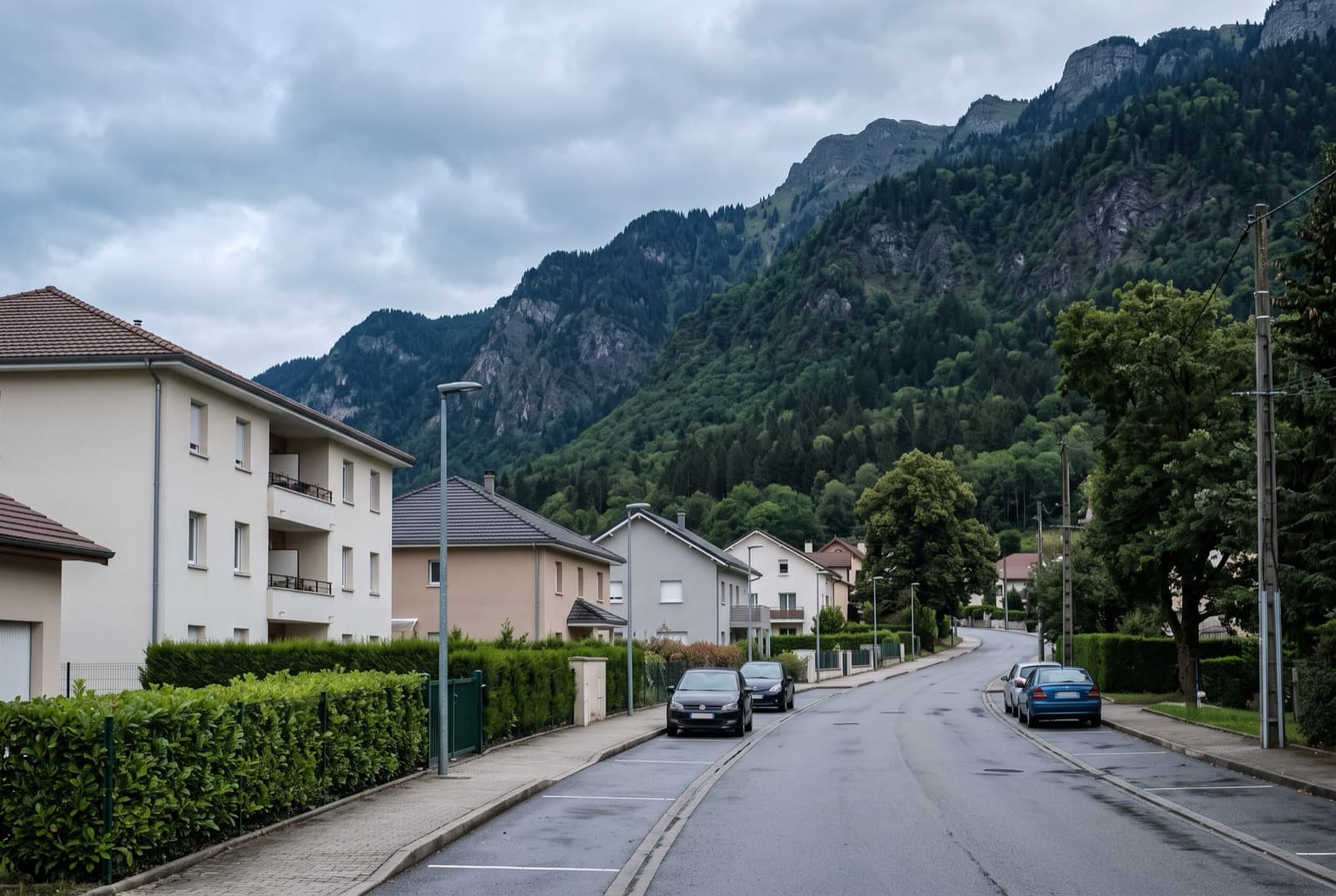 Rue de commune périurbaine avec maisons pavillonnaires et petits immeubles en enduit clair, versant boisé en fond